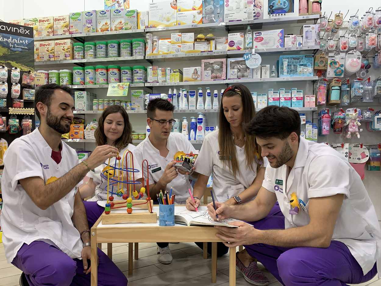 Una farmac&eacute;utica en primer plano con los brazos cruzados sonr&iacute;e mientras dos farmac&eacute;uticos en el fondo charlan